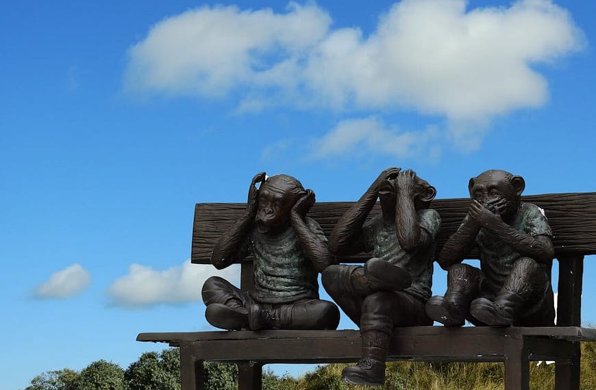 Artistic sculpture depicting the Three Wise Monkeys on a bench outdoors against blue sky.